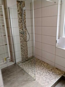 Glass shower enclosure with pebble mosaic floor and vertical pebble strip in a tiled bathroom; handheld showerhead.