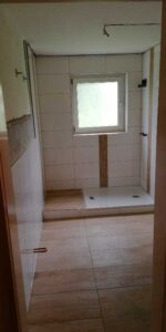 Bathroom under construction: white tiled shower area with a small window, shower tray, and exposed plumbing and fixtures awaiting installation.