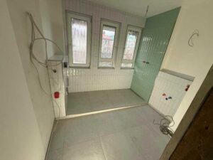 Unfinished bathroom with white tile walls, three small windows, and a green glass shower area; exposed pipes and red outlet.