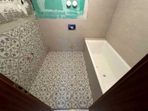 Bathroom under renovation with patterned gray-and-white floor tiles, a white rectangular bathtub along the right wall, and exposed plumbing fixtures. A green drywall patch and unfinished walls indicate ongoing construction.