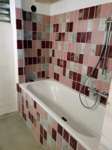 Pink, gray, and maroon tile surround around a white built-in bathtub, with a handheld showerhead and chrome fixtures nearby.