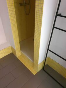 Shower alcove with yellow mosaic tiles on walls and floor, and a handheld shower head mounted inside.