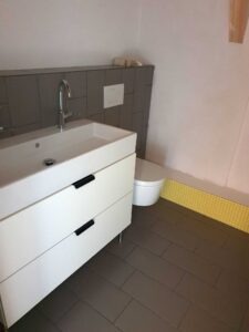 Modern bathroom with a white vanity sink, chrome faucet, and grey tiled backsplash; wall-mounted dual-flush plate behind the sink; small wooden block on a ledge above; white toilet to the right; yellow tiled trim along the baseboard.