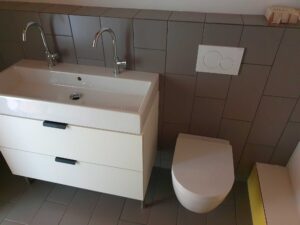 White bathroom vanity with two chrome faucets and a rectangular sink, next to a wall with gray tiles and a dual flush plate on the wall behind the toilet