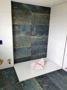 Under-construction bathroom shower area with dark slate tiles on the wall and a white bench; exposed plumbing fittings nearby.