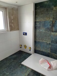 Under‑construction bathroom with white tiled walls, a tall white cabinet, and dark green slate tile on the adjacent wall; exposed yellow plumbing fittings near the floor.