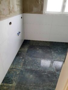 Bathroom in progress with white wall tiles installed, exposed concrete wall above, and a window to the right.