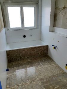 Under-construction bathroom with a built-in bathtub, white wall tiles, and a large window; exposed plumbing fittings and unfinished floor.