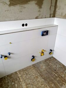 Under-construction kitchen wall with white cabinets, exposed plumbing connections, and open circular pipe holes above a tiled floor