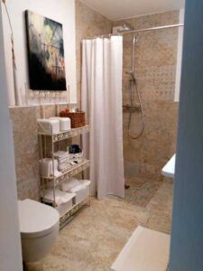 Small tiled bathroom with a walk-in shower behind a white curtain, a metal shelf stacked with towels, and a wall-mounted toilet.