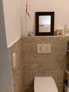 Bathroom with beige tiled half-wall, white dual-flush plate, and a white toilet below. A small mirror sits on the shelf above, with decorative items nearby.