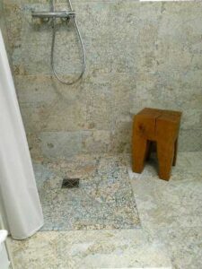 Shower area with handheld showerhead, tiled walls, and a wooden stool beside the drain area in a light-colored stone tile room