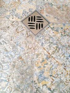 Decorative tiled floor with blue, gold, and white floral patterns; a square metal drain cover with slotted openings near the top center.