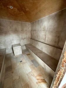 Tiled sauna with a long bench along the wall and a small square stool; beige stone tiles throughout.