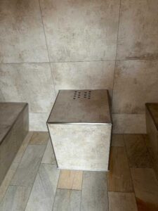Rectangular metal trash bin with a perforated top, standing against tiled walls in a tiled room.