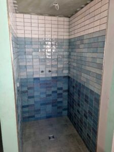 Small shower stall with blue gradient tiles from dark floor to lighter walls, floor drain visible, ceiling opening with pipe, walls fully tiled