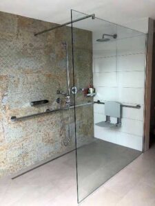Accessible glass-walled shower with beige stone and white tile walls, multiple grab bars, a handheld shower, and a recessed shelf for toiletries.