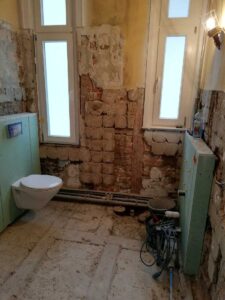 Bathroom under renovation with exposed brick walls, a wall-mounted toilet, and exposed pipes on the floor surrounded by tools and debris.