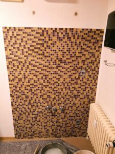 Bathroom wall covered in small yellow-brown mosaic tiles with exposed faucets and a radiator to the right; renovation tools visible on the floor in the foreground.