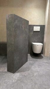Wall-mounted toilet with dual-flush plate visible on the tiled wall, next to a dark stone partition in a modern restroom.