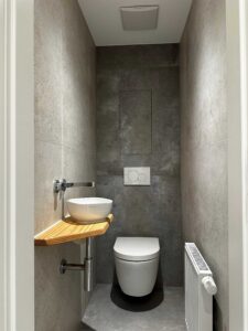Narrow modern bathroom with gray textured walls, white vessel sink on a wooden shelf, chrome faucet, and a wall-mounted dual-flush plate above the toilet.