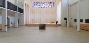 Interior of a bright, minimalist chapel with a central perforated metal altar, vertical wooden slats wall, and colorful stained-glass window above.