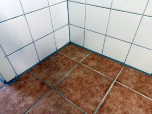 Corner of a bathroom where white wall tiles meet brown floor tiles with visible grout lines in a diagonal perspective.