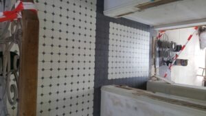 White tiled floor with white tiles and small dark diamond accents, bordered by a dark tile stripe in a construction area.