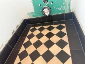Checkerboard beige and black tile floor in a small unfinished room with exposed plumbing on a green wall patch and dark base tiles around the edge.