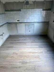 Room under renovation with white square tile backsplash, exposed plumbing, and unfinished concrete walls; new wood-look flooring being installed.