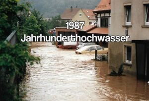 '1987 Jahrhunderthochwasser' text over a flooded German town street with cars partially submerged and water reaching doorways.